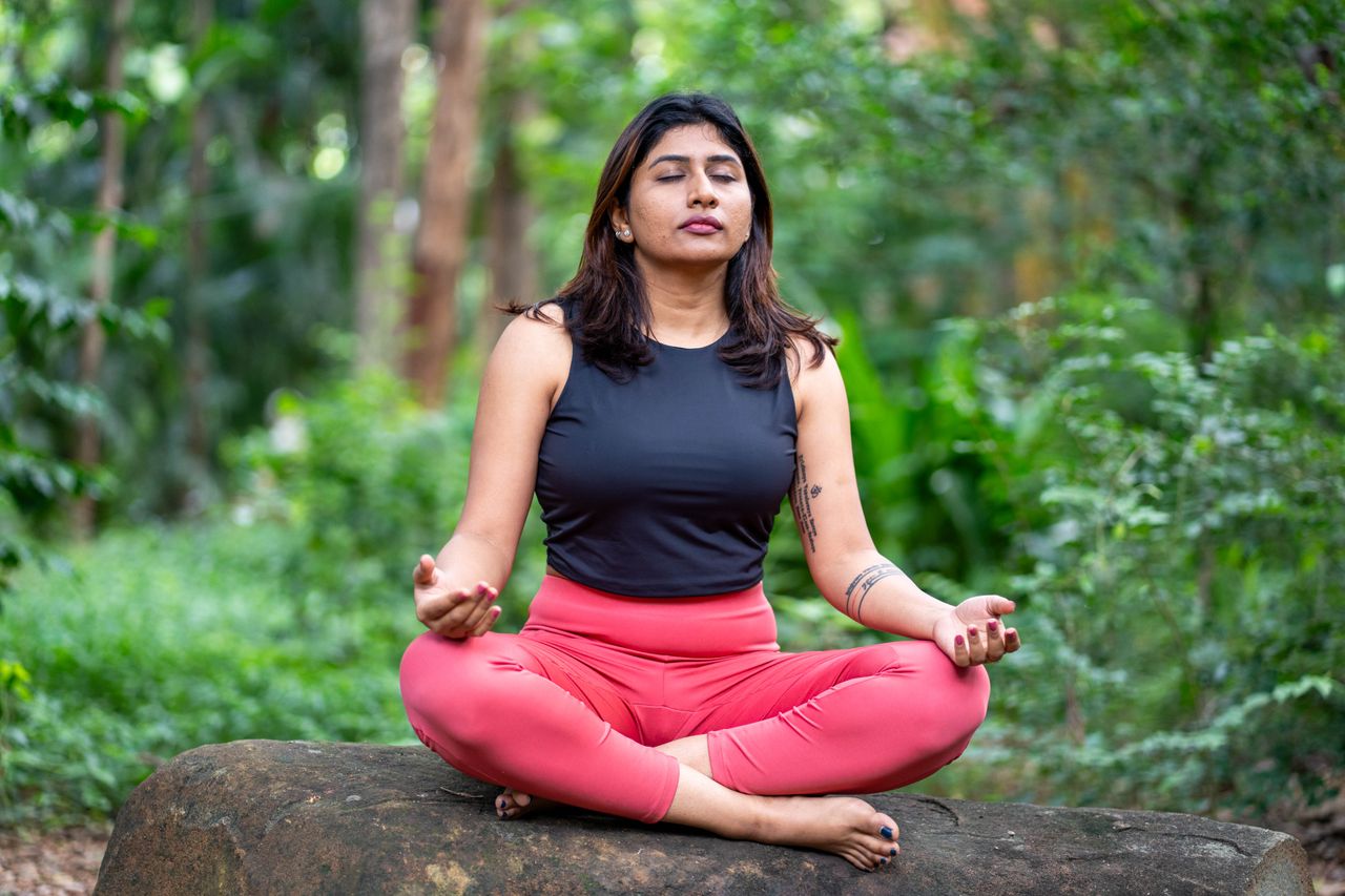 A woman practicing meditation outdoors surrounded by lush greenery.